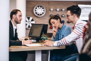 Conseiller automobile expliquant les modalités de financement et de TVA à un jeune couple dans une concession