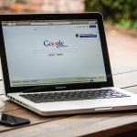 Laptop ouvert sur Google, smartphone à gauche, table en bois, jardin flou